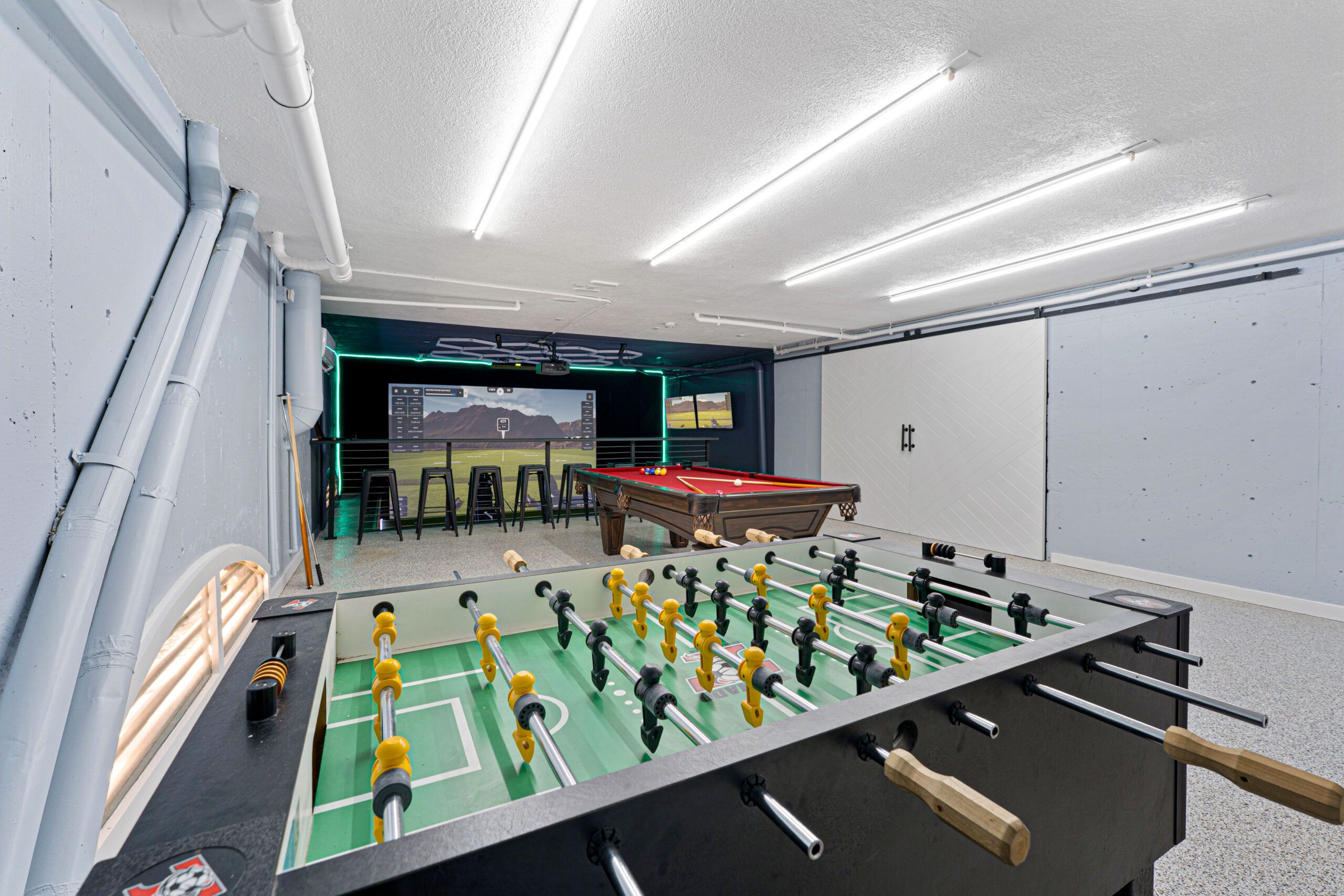 Wide game room with a foosball table, red-felt billiards, and the golf simulator screen at the estate